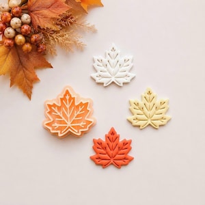 May include: A set of four maple leaf-shaped items including one orange plastic cookie cutter and three pressed leaf shapes in white, pale yellow, and burnt orange. The items are arranged near a cluster of artificial autumn leaves and berries.