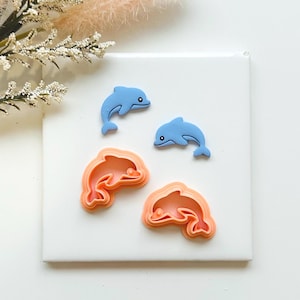 May include: Four dolphin-themed items on a white surface. Two light blue dolphin figurines and two peach-coloured dolphin-shaped cookie cutters are arranged on a square tile. Decorative floral elements are in the background.