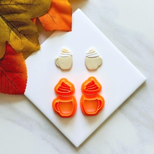 May include: Two sets of coffee-themed polymer clay cutters and finished earrings. The cutters are bright orange, shaped like mugs with whipped cream. The earrings are beige mugs with white whipped cream. Autumn leaves in orange and yellow are in the background.