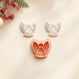 May include: Two white angel-shaped ornaments with gold fleck details and one peach-colored plastic angel-shaped cookie cutter. The ornaments feature a circular head and wings with etched lines, while the cutter shows the internal structure.
