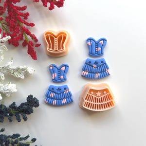 May include: A collection of cookie cutters in the shape of a corset and skirt. The corset cutters are peach-colored, while the skirt cutters are blue with pink accents. The cutters are arranged on a white surface.