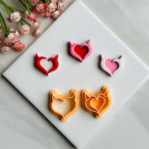Op de afbeelding: Vier hartvormige koekjesvormen met duivelshoorns. Twee zijn rood, één is roze en één is oranje.