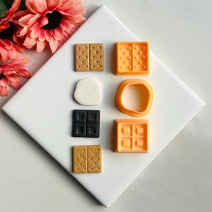 K&ouml;nnte beinhalten: Ein Set mit sechs Miniatur-Zutaten f&uuml;r S'mores, darunter Graham-Cracker, Schokoladenquadrate und Marshmallows. Die Artikel sind aus Polymer Clay gefertigt und perfekt zum Basteln oder Dekorieren.