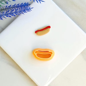 May include: Two miniature hot dog charms, one with a red ketchup topping and the other with a yellow mustard topping. The charms are on a white background.