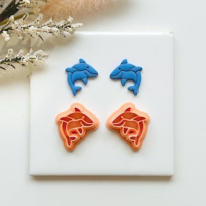 May include: Four dolphin-shaped craft supplies in blue and orange. Two blue dolphins are on top, and two orange dolphins are below. The dolphins are on a white surface, with a sprig of white flowers in the background.