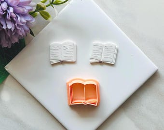 May include: Three open book-shaped cookie cutters. Two are white, and one is peach-colored. The cutters are arranged on a white surface, with a purple flower and green leaves in the background. The cutters are for baking.