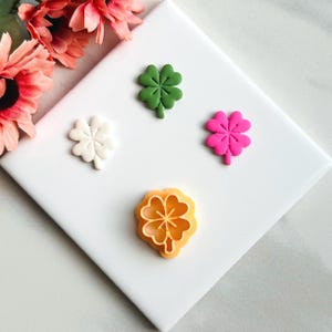 Puede incluir: Decoraciones de tréboles de cuatro hojas en verde, blanco, rosa y naranja. Los tréboles están dispuestos sobre una superficie blanca, con algunas flores rosas en el fondo. Ideal para el Día de San Patricio o cualquier ocasión.