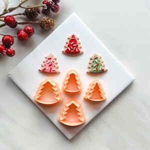 May include: A collection of Christmas tree-shaped cookie cutters and decorated cookies. The cookies are frosted in red, pink, and green, with colorful sprinkles. The cutters are made of orange plastic. The image is set on a white surface with decorative berries.