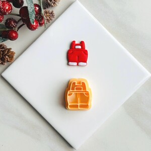 May include: A red and white fondant cookie cutter and a red fondant cookie in the shape of dungarees.