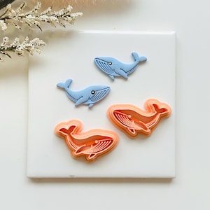 May include: Three whale-shaped items. Two are light blue, with detailed lines and a small eye. The other two are orange, with layered outlines. All are on a white surface, with a sprig of white flowers in the upper left corner.