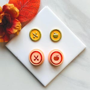 Könnte beinhalten: Vier runde, dekorative Stempel mit geprägten Designs. Zwei sind gelb mit Scheren- und Apfelmotiven, und zwei sind pfirsichfarben mit Scheren- und Apfelmotiven. Die Stempel sind auf einer weißen Oberfläche mit einem herbstlichen Blatt und einer Blume angeordnet.