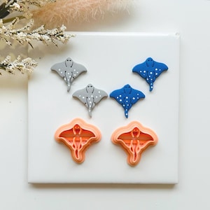 May include: A selection of handmade stingray-shaped items. The image displays grey and blue stingray figurines with white polka dots, alongside two orange cookie cutters in the same shape. The items are arranged on a white surface.