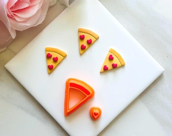 Könnte beinhalten: Eine Sammlung von Bastelmaterialien zum Thema Pizza. Das Bild zeigt vier Pizza-Scheiben-Anhänger mit roten Herz-Toppings, einen orangefarbenen Pizzaschneider und einen kleinen herzförmigen Anhänger. Die Pizzen haben gelbe Krusten und liegen auf einer weißen Oberfläche.