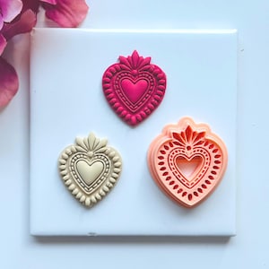 Puede incluir: Tres cortadores de galletas en forma de corazón en tonos rosa y crema. El cortador superior es rosa intenso, el del medio es crema y el inferior es rosa claro. Cada cortador tiene un diseño de corazón detallado con bordes decorativos.