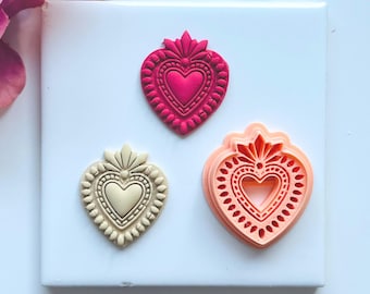 Könnte beinhalten: Drei herzförmige Ausstecher in Rosa- und Cremetönen. Der obere Ausstecher ist tiefrosa, der mittlere cremefarben und der untere hellrosa. Jeder Ausstecher hat ein detailliertes Herzdesign mit dekorativen Rändern.