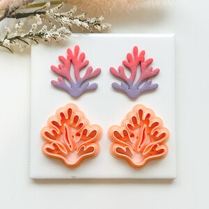 May include: Four coral-shaped polymer clay earrings. Two pairs of earrings are displayed on a white square tile. The top pair is coral and purple, and the bottom pair is orange. The earrings are designed to resemble coral.
