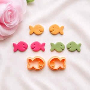 May include: A collection of small fish-shaped items including two orange solid fish, two pink solid fish, two light green solid fish, and two orange fish-shaped cookie cutters with open centres, all arranged on a soft white fabric surface.