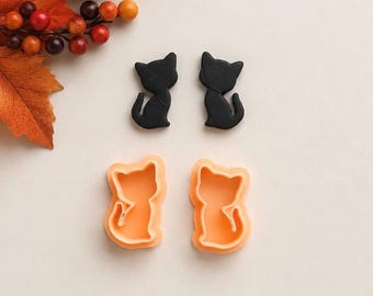 May include: Two black cat-shaped cookie cutters and two orange cat-shaped cookie cutters arranged in a square formation. The black cutters are positioned above the orange cutters, each featuring a silhouette of a sitting cat with a curved tail.