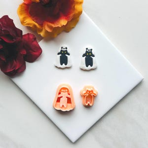 Puede incluir: Una colección de suministros para manualidades con temática de Halloween. Dos decoraciones blancas en forma de fantasma con cuerpos negros y ojos amarillos se encuentran encima de dos cortadores de galletas naranjas. Una flor roja y amarilla está en el fondo.