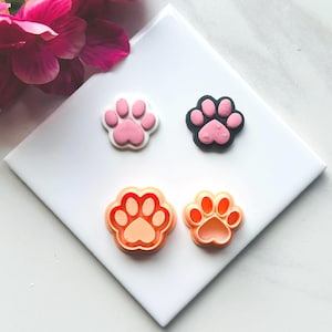 May include: Four paw print shaped cookie cutters, two are orange, one is pink, and one is black. The cutters are on a white surface.
