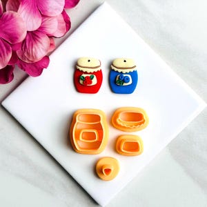Puede incluir: Un juego de cinco cortadores de arcilla polimérica para hacer tarros de mermelada en miniatura. Los cortadores tienen la forma de un tarro, una tapa y dos círculos de diferentes tamaños. El juego incluye dos tarros de mermelada de arcilla polimérica terminados, uno rojo con bayas rojas y uno azul con bayas azules.