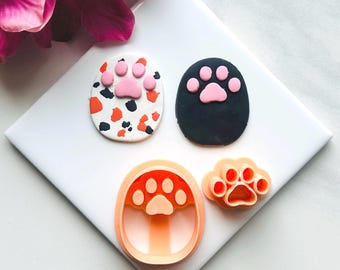 Puede incluir: Tres cortadores de galletas de plástico naranja en forma de huella de pata. Dos de los cortadores son más grandes y tienen un diseño de huella de pata con un centro rosa. El tercer cortador es más pequeño y tiene un diseño de huella de pata con un centro rosa.