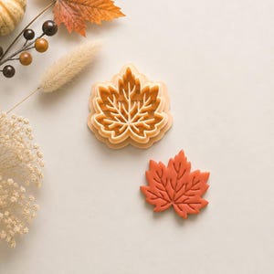 Puede incluir: Un cortador de galletas con forma de hoja de arce en color naranja claro y una galleta de hoja de arce naranja a juego. El cortador presenta un patrón de venas detallado para marcar la masa, mientras que la galleta muestra la forma.