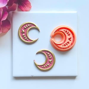 May include: Three crescent moon-shaped cookie cutters. Two are pink with gold leaf accents, and one is orange. The cutters are on a white surface with a pink flower in the background. The cutters are for baking.