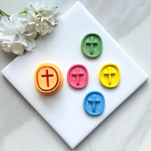May include: Five colorful cross-shaped cookie cutters, including yellow, pink, blue, green, and orange. The cutters are on a white surface.