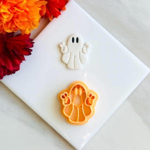 May include: Two Halloween-themed cookie cutters and a fondant ghost. The white fondant ghost is in the shape of a friendly ghost with two black eyes and two hands making peace signs. The orange cookie cutter is in the shape of a ghost.
