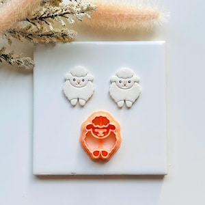 May include: Three white sheep-shaped decorations with black eyes and orange cookie cutter. The cookie cutter is orange and has the same sheep shape. The decorations are on a white surface.