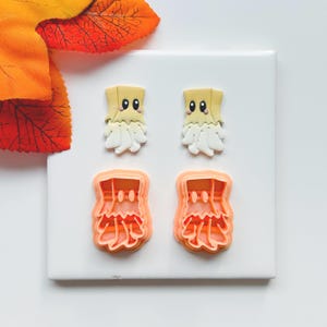 May include: Two sets of Halloween-themed cookie cutters and two finished cookies. The cookies are shaped like ghosts with beige heads, white bodies, and black eyes. The cutters are orange and have the same ghost shape.