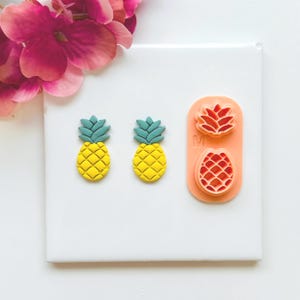 May include: Two yellow and green pineapple-shaped earrings and two pineapple-shaped cookie cutters on a white surface. The cookie cutters are in a peach-colored holder. Pink flowers are in the background.