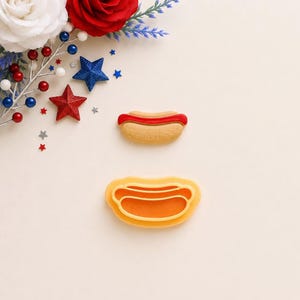 May include: A tan and red hot dog shaped cookie cutter and a matching tan and red hot dog shaped cookie. The cookie cutter is made of orange plastic and the cookie features a textured tan bun with a smooth red line representing the hot dog.