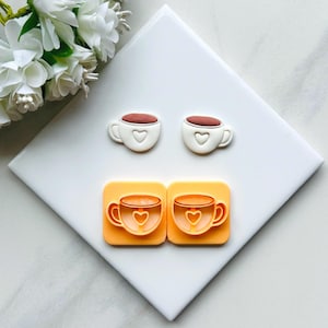 May include: Two white ceramic coffee cups with a heart design and two orange plastic moulds of coffee cups with a heart design.