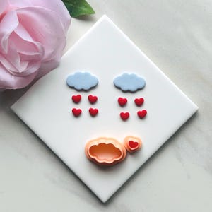 Puede incluir: Una baldosa cuadrada blanca presenta un diseño de dos nubes azul claro con gotas de lluvia en forma de corazón rojo. Debajo, hay dos cortadores de galletas, uno en forma de nube y otro en forma de corazón. Una flor rosa está en el fondo.