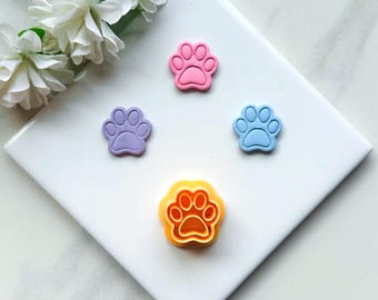 May include: Four paw print cookie cutters, one each in pink, purple, blue, and yellow. The cutters are on a white surface.