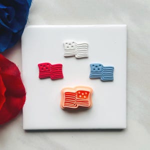 Puede incluir: Cuatro pequeños adornos con forma de bandera en rojo, blanco y azul, dispuestos sobre una baldosa cuadrada blanca. Las banderas tienen un diseño ondulado con estrellas en la esquina superior izquierda. Una flor roja y azul está en el fondo.