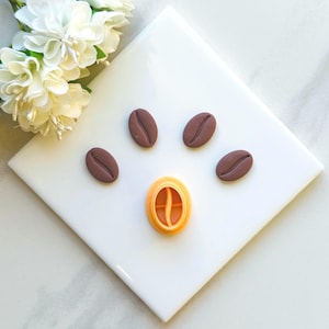 May include: Five brown coffee bean shaped fondant decorations and a yellow fondant coffee bean cutter on a white surface.