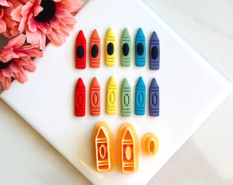 May include: A collection of colorful crayon-shaped objects arranged on a white surface. The crayons are in various colors, including red, orange, yellow, green, blue, and purple. Also included are orange crayon-shaped molds and a small ring.