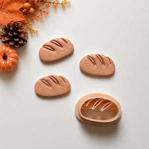 Peut inclure: Trois découpes de pain ovales, texturées et brun clair, accompagnées d'un emporte-pièce en plastique assorti. Les découpes présentent trois lignes diagonales sur la surface supérieure, imitant l'aspect d'une miche de pain croustillante.
