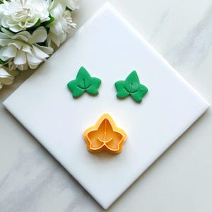 May include: Two green plastic leaf-shaped cookie cutters and one orange plastic leaf-shaped cookie cutter on a white surface.