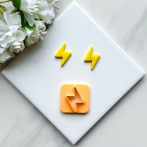 Può includere: Due orecchini a forma di fulmine gialli e un tagliapasta a forma di fulmine giallo su una superficie bianca.