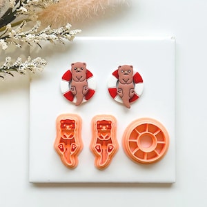 May include: A selection of otter-themed cookie cutters and decorations. Two brown otters are in red and white life preservers. There are also three orange cookie cutters in the shape of otters and a round cutter.