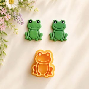 May include: Two green frog-shaped cookie cutters with black eyes and a smiling mouth, positioned above one orange frog-shaped cookie cutter. The cutters feature a raised outline design of a seated frog with front legs and hind legs clearly defined.