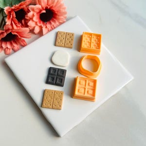 K&ouml;nnte beinhalten: Ein Set mit sechs Miniatur-Formen aus Silikon in Lebensmittelform zum Basteln. Die Formen haben die Form von Graham-Crackern, Schokoladenquadraten, Marshmallows und Waffelquadraten. Die Formen sind orange und braun.