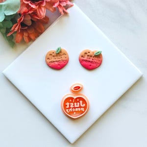 May include: Three peach-shaped earrings with the text "Just Peachy" in a pink and orange gradient.