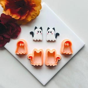 May include: A collection of Halloween-themed craft supplies. Two white ghost cat earrings with black accents and two orange ghost cookie cutters are displayed with two orange cat cookie cutters. A white surface is the backdrop.
