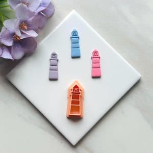 Op de afbeelding: Vier gekleurde objecten in de vorm van een vuurtoren in paars, blauw, roze en oranje. De objecten zijn gerangschikt op een witte vierkante tegel, met een paarse bloem in de linkerbovenhoek. De vuurtorens hebben een eenvoudig, gestileerd ontwerp.