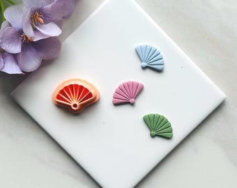 May include: A collection of colorful fan-shaped objects in orange, pink, blue, and green, arranged on a white square tile. The orange fan has a detailed interior design. The other fans are solid colors.